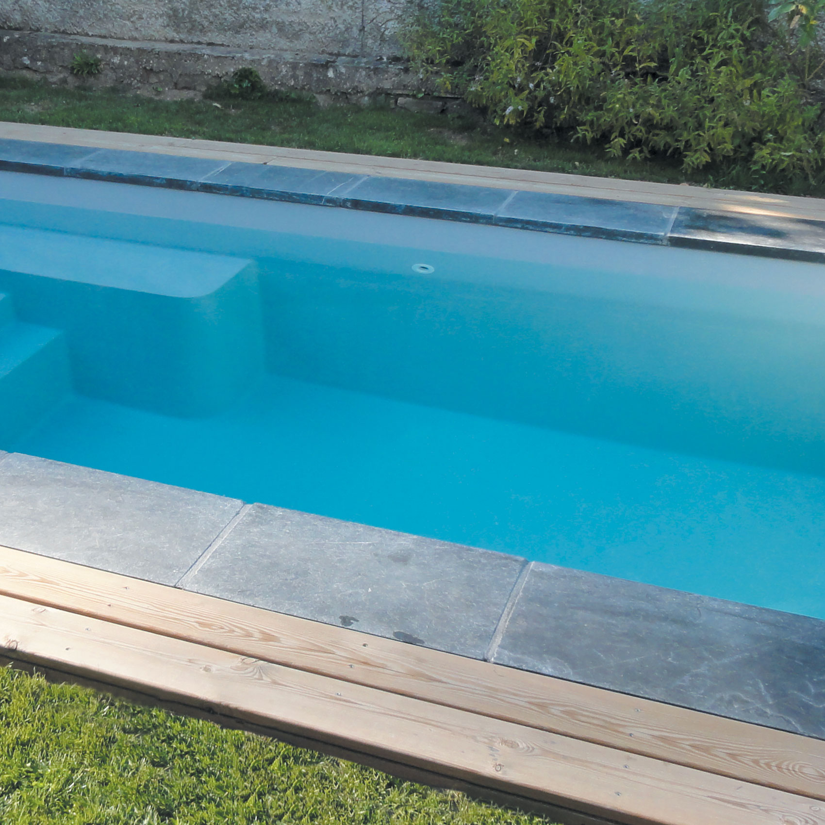 Piscine rectangulaire moderne avec margelle en pierre et entourage en bois dans un jardin verdoyant.