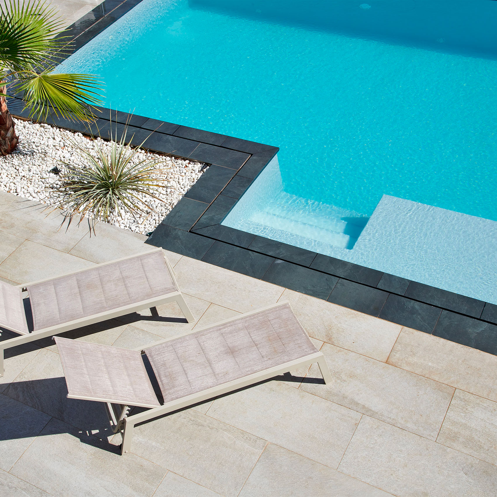 Vue aérienne d'une piscine bleue avec deux chaises longues et décoration contemporaine avec plantes et galets.