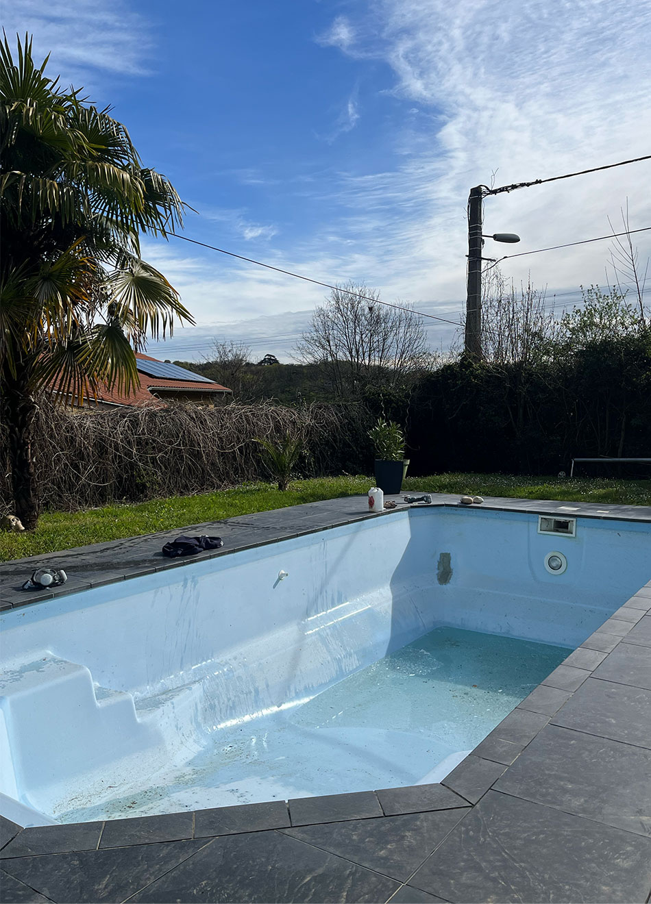 Piscine extérieure vide sous un ciel bleu avec des plantes et un poteau électrique.